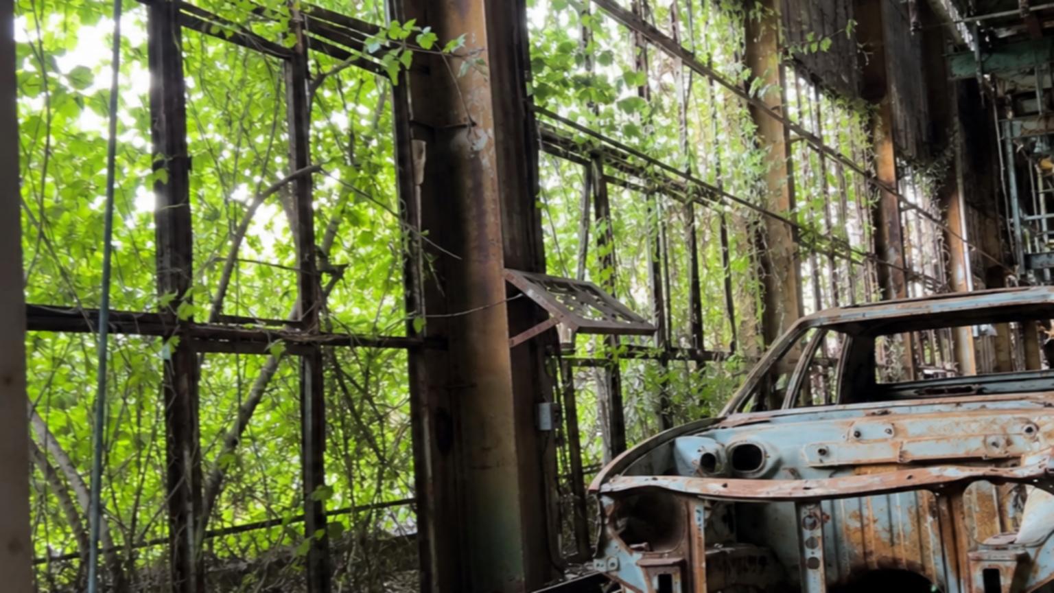An abandoned, rusting car factory with vines creeping through broke…